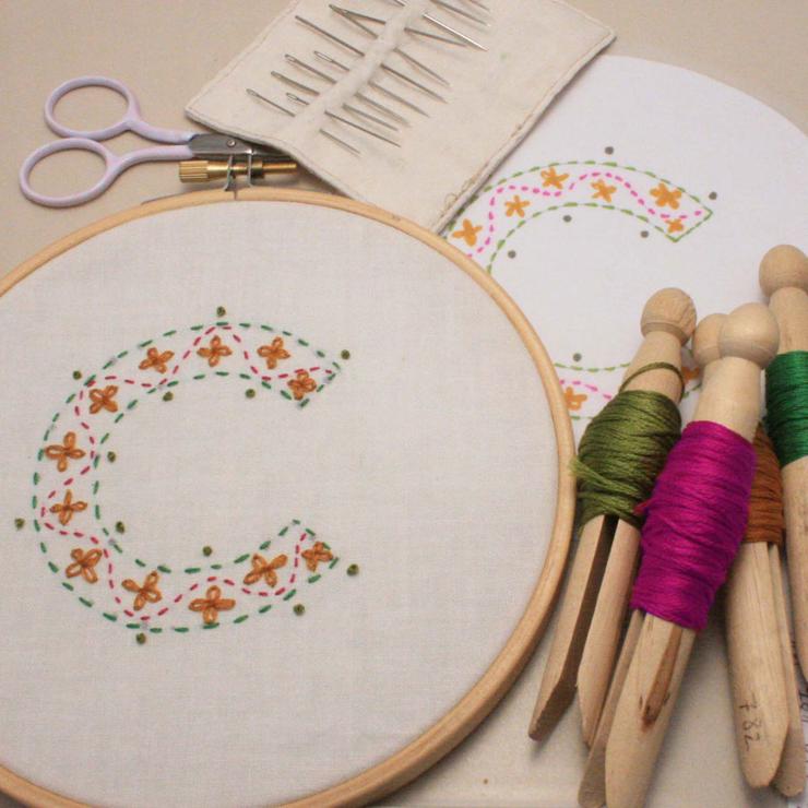 hand embroidered letter C in shades of mustard yellow  and fuschia pink with bobbins of thread arranged beside the embroidery hoop.